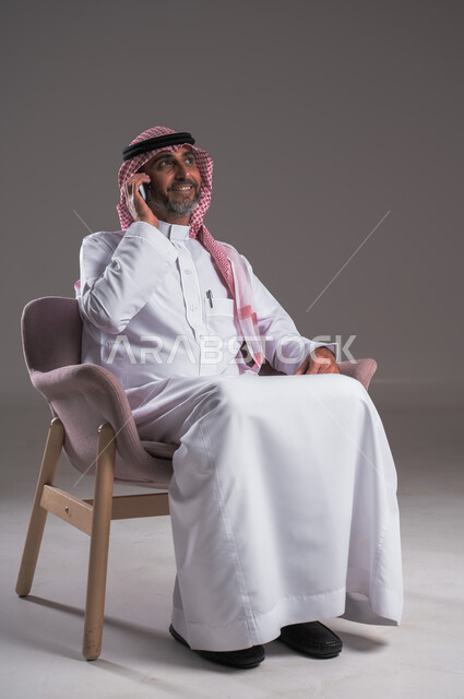 إجراء مكالمات هاتفية عبر الهاتف المحمول, التواصل مع الأهل والأصدقاء، بورتريه لرجل عربي سعودي خليجي مبتسم يرتدي الثوب التقليدي والشماغ يجلس على كرسي مريح ويستخدم الجوال، استخدام الوسائل التقنية الحديثة في الحياة العملية، الشعور بالسعادة لسماع خبر ما، خلفية رمادية
