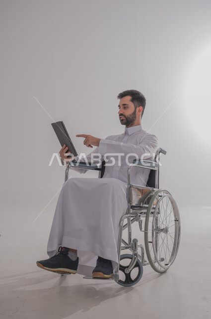 إجراء مكالمات الفيديو باستخدام جهاز التابلت، الاستفادة من التقنيات الحديثة والتكنولوجيا المتطورة، بورتريه لشاب عربي خليجي سعودي من ذوي الاحتياجات الخاصة يرتدي الثوب التقليدي يجلس على كرسي متحرك يتواصل مع الأهل والأصدقاء بإيماءات الاندماج، خلفية رمادية