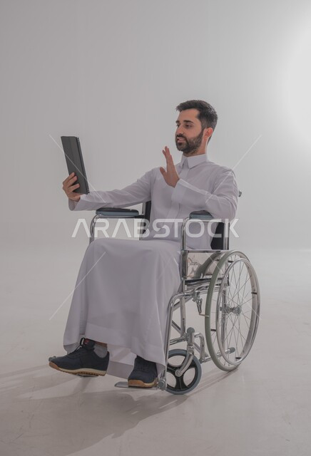 إجراء مكالمات الفيديو باستخدام جهاز التابلت، الاستفادة من التقنيات الحديثة والتكنولوجيا المتطورة، بورتريه لشاب عربي خليجي سعودي من ذوي الاحتياجات الخاصة يرتدي الثوب التقليدي يجلس على كرسي متحرك يتواصل مع الأهل والأصدقاء بإيماءات الاندماج، خلفية رمادية