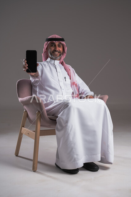 النظر إلى الكاميرا بايماءات الفرح والسرور، بورتريه لرجل عربي سعودي خليجي مبتسم يرتدي الثوب التقليدي والشماغ يجلس على كرسي مريح ويحمل الجوال، عرض شاشة سوداء فارغة على الهاتف المحمول، استخدام الوسائل التقنية الحديثة المتطورة، خلفية رمادية