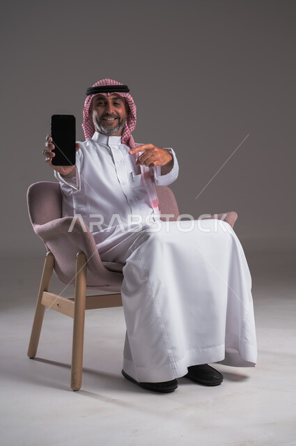 عرض شاشة سوداء فارغة على الهاتف المحمول، استخدام الوسائل التقنية الحديثة، النظر إلى الكاميرا بايماءات الفرح والسرور، بورتريه لرجل عربي سعودي خليجي مبتسم يرتدي الثوب التقليدي والشماغ يجلس على كرسي مريح ويحمل الجوال ويشير اليه، خلفية رمادية