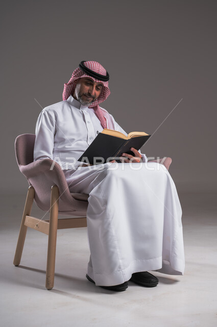 تمضية أوقات الفراغ بالتعلم والتثقف، الاهتمام بتصفح الكتب المفيدة، حب المطالعة والقراءة، بورتريه لرجل عربي خليجي سعودي يرتدي الثوب التقليدي والشماغ يجلس على كرسي مريح ينظر الى الكتاب بتمعن وتركيز، ثقافة التعليم الذاتي، خلفية رمادية