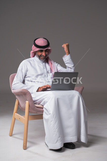تعابير الفوز والنصر وتحقيق أهداف العمل الاداري, ايماءات وجه ويدين تدل على السرور والبشاشة وإنجاز المهام المطلوبة, الحماس والقيام بصفقات ناجحة مع العملاء عبر اللابتوب, بورتريه لرجل عربي خليجي سعودي يرتدي الثوب التقليدي والشماغ يجلس على كرسي مريح، بخلفية رمادية