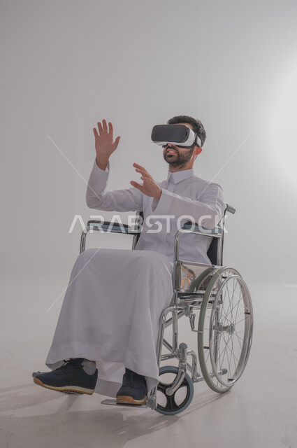 Merging with virtual reality, taking advantage of modern techniques and advanced technology, portrait of a young Saudi Arabian Gulf man with special needs wearing traditional dress sitting on a wheelchair wearing VR glasses and raising his hand with gestures of interaction with imaginary elements, gray background