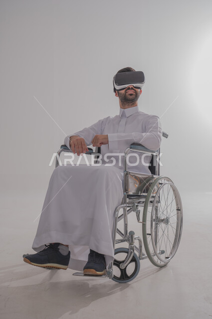 Merging with virtual reality, taking advantage of modern techniques and advanced technology, portrait of a young Saudi Arabian Gulf man with special needs wearing traditional dress sitting on a wheelchair wearing VR glasses and raising his head with gestures interacting with imaginary elements, gray background