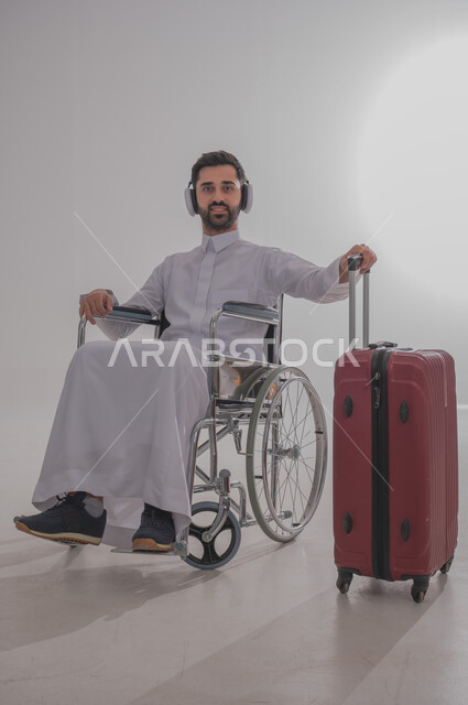 Preparing to go on exciting recreational trips, portrait of a young Saudi Arabian Gulf man with special needs wearing traditional dress sitting on a wheelchair, putting on headphones and listening to his favorite music and songs, holding a red suitcase and preparing to leave, gray background