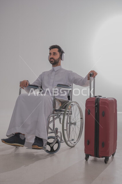 الاستعداد للذهاب في رحلات ترفيهية مشوقة، بورتريه لشاب عربي خليجي سعودي من ذوي الاحتياجات الخاصة يرتدي الثوب التقليدي يجلس على كرسي متحرك يضع سماعات الهيدفون ويستمع للموسيقا والأغاني المفضلة لديه، الإمساك بحقيبة السفر الحمراء والتجهيز للرحيل، خلفية رمادية