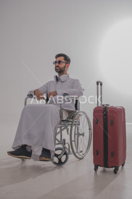 الاستعداد للذهاب في رحلات ترفيهية مشوقة، بورتريه لشاب عربي خليجي سعودي من ذوي الاحتياجات الخاصة يرتدي الثوب التقليدي يجلس على كرسي متحرك يضع نظارة شمسية وسماعات الهيدفون ويستمع للموسيقا المفضلة لديه، تجهيز حقيبة السفر الحمراء من أجل الرحيل، خلفية رمادية