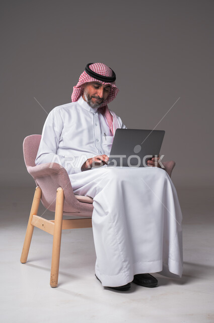 استخدام الحاسوب المحمول لإنجاز المهام، بورتريه لرجل عربي سعودي خليجي يرتدي الثوب التقليدي والشماغ يجلس عل كرسي مريح ويحمل جهاز اللابتوب، تطور التكنولوجيا والأجهزة التقنية، تصفح برامج التواصل الاجتماعي, عقد إجتماع عمل عبر الإنترنت، خلفية رمادية