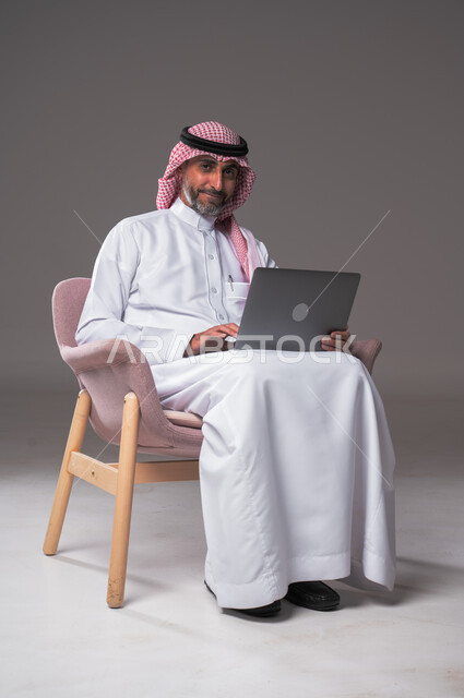 عقد إجتماع عمل عبر الإنترنت، إستخدام الحاسوب المحمول لإنجاز المهام، بورتريه لرجل عربي سعودي خليجي يرتدي الثوب التقليدي والشماغ يجلس عل كرسي مريح ويحمل جهاز اللابتوب، تطور التكنولوجيا والأجهزة التقنية، تصفح برامج التواصل الاجتماعي, خلفية رمادية