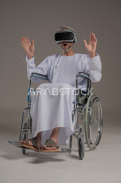  تحسين جودة حياة ذوي الاعاقة الحركية، استخدام كراسي المعاقين للمساعدة على الحركة والتنقل، بورتريه لرجل عربي  خليجي عماني يرتدي العمامة والدشداشة يقوم بمشاهدة أفلام عبر نظارات الواقع الافتراضي ثلاثية الأبعاد، محاكاة العالم الخيالي والتفاعل مع الفيلم، خلفية رمادية