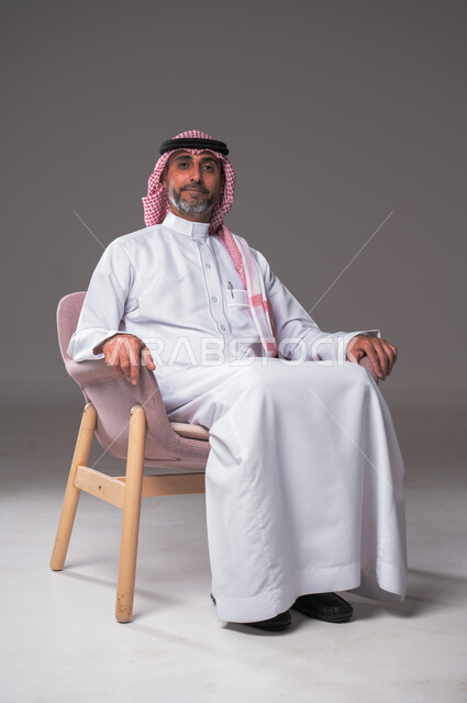 مفهوم الوسامة والرجولة، بورتريه لرجل عربي سعودي خليجي يرتدي الثوب التقليدي والشماغ يجلس عل كرسي مريح، النظر إلى الكاميرا بإيماءات الثقة بالنفس، الاهتمام بالأناقة والمظهر الخارجي، خلفية رمادية