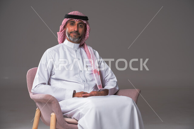الاهتمام بالأناقة والمظهر الخارجي، مفهوم الوسامة والرجولة، بورتريه لرجل عربي سعودي خليجي يرتدي الثوب التقليدي والشماغ يجلس عل كرسي مريح، النظر إلى الكاميرا بإيماءات الثقة بالنفس، خلفية رمادية