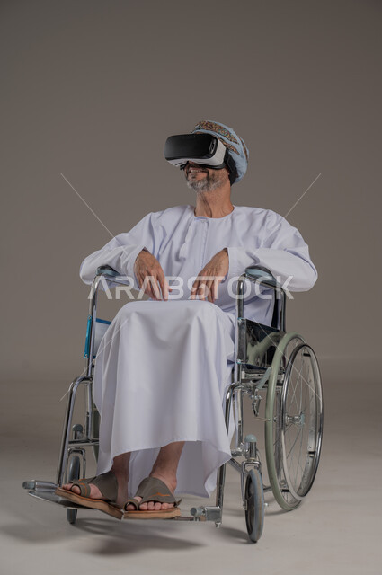 محاكاة العالم الخيالي والتفاعل مع الفيلم، تحسين جودة حياة ذوي الاعاقة الحركية، استخدام كراسي المعاقين للمساعدة على الحركة والتنقل، بورتريه لرجل عربي  خليجي عماني يرتدي العمامة والدشداشة يقوم بمشاهدة أفلام عبر نظارات الواقع الافتراضي ثلاثية الأبعاد، خلفية رمادية