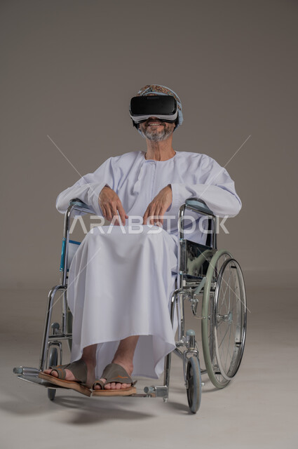محاكاة العالم الخيالي والتفاعل مع الفيلم، تحسين جودة حياة ذوي الاعاقة الحركية، استخدام كراسي المعاقين للمساعدة على الحركة والتنقل، بورتريه لرجل عربي  خليجي عماني يرتدي العمامة والدشداشة يقوم بمشاهدة أفلام عبر نظارات الواقع الافتراضي ثلاثية الأبعاد، خلفية رمادية