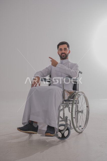 ضعف القدرة الحركية وصعوبة المشي، بورتريه لشاب عربي خليجي سعودي من ذوي الاحتياجات الخاصة يرتدي الثوب التقليدي يجلس على كرسي متحرك يشير باصبع يده إلى مكان ما، النظر للكاميرا بإيماءات البشاشة والسرور، الجسم بالطول الكامل، خلفية رمادية