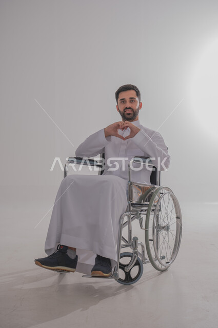 ضعف القدرة الحركية وصعوبة المشي، بورتريه لشاب عربي خليجي سعودي من ذوي الاحتياجات الخاصة يرتدي الثوب التقليدي يجلس على كرسي متحرك يصنع بيده شكل قلب بتعبيرات المحبة والمودة، النظر للكاميرا بإيماءات البشاشة والسرور، الجسم بالطول الكامل، خلفية رمادية