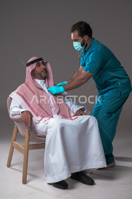 استخدام سماعة الطبيب لسماع نبضات القلب، مفهوم الطب والرعاية الصحية، بورتريه لممرض يرتدي اليونيفورم والقفازات والكمامة الوقائية يقوم بفحص رجل مسن عربي خليجي سعودي يلبس الثوب التقليدي والشماغ يجلس على كرسي مريح، تقديم الخدمات الصحية للمرضى، خلفية رمادية