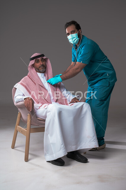 استخدام سماعة الطبيب لسماع نبضات القلب، مفهوم الطب والرعاية الصحية، بورتريه لممرض يرتدي اليونيفورم والقفازات والكمامة الوقائية يقوم بفحص رجل مسن عربي خليجي سعودي يلبس الثوب التقليدي والشماغ يجلس على كرسي مريح، تقديم الخدمات الصحية للمرضى، خلفية رمادية