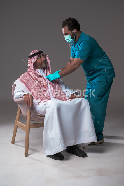 استخدام سماعة الطبيب لسماع نبضات القلب، مفهوم الطب والرعاية الصحية، بورتريه لممرض يرتدي اليونيفورم والقفازات والكمامة الوقائية يقوم بفحص رجل مسن عربي خليجي سعودي يلبس الثوب التقليدي والشماغ يجلس على كرسي مريح، تقديم الخدمات الصحية للمرضى، خلفية رمادية