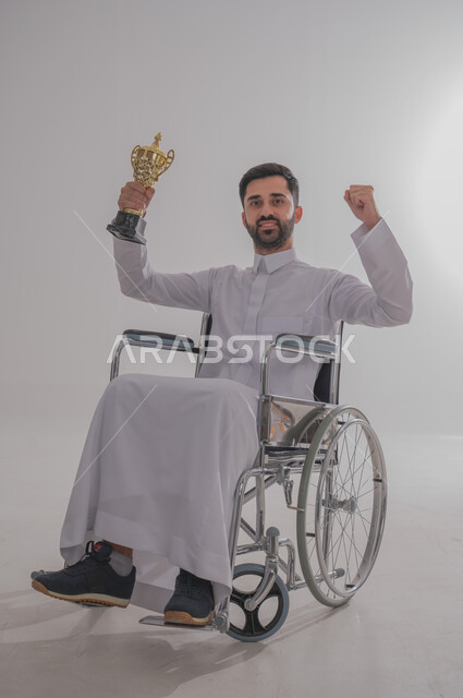 التتويج بالبطولة بعد الفوز والحصول على المركز الأول، بورتريه لشاب عربي خليجي سعودي من ذوي الاحتياجات الخاصة يرتدي الثوب التقليدي يجلس على كرسي متحرك يحمل الكأس الذهبي ويرفع يديه بحماس، النظر للكاميرا بإيماءات البشاشة والسرور، الجسم بالطول الكامل، خلفية رمادية