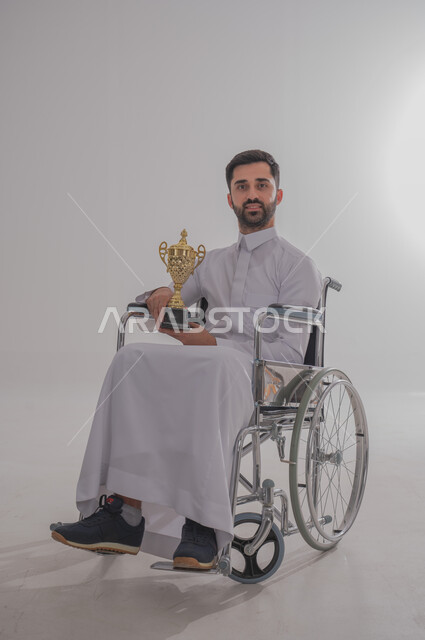 التتويج بالبطولة بعد الفوز والحصول على المركز الأول، بورتريه لشاب عربي خليجي سعودي من ذوي الاحتياجات الخاصة يرتدي الثوب التقليدي يجلس على كرسي متحرك يحمل بيده الكأس الذهبي بحماس، النظر للكاميرا بإيماءات البشاشة والسرور، الجسم بالطول الكامل، خلفية رمادية