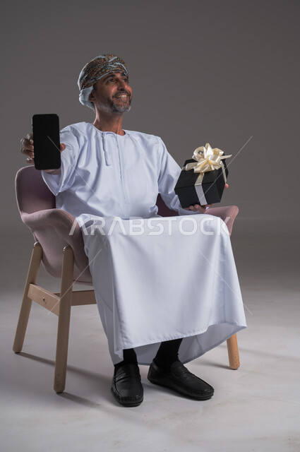  دعم المجتمع لذوي الهمم, عرض شاشة سوداء فارغة، مناسبات و أعياد سعيدة، بورتريه لرجل عربي  خليجي عماني يرتدي العمامة والدشداشة يجلس على كرسي متحرك يحمل بيده صندوق هدايا والهاتف المحمول، شعور المعاقين بالحب و التقدير، صورة لكامل الجسم، خلفية رمادية