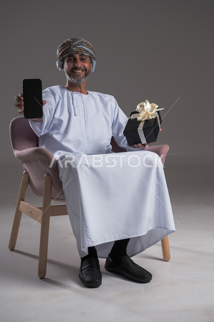  دعم المجتمع لذوي الهمم, عرض شاشة سوداء فارغة، مناسبات و أعياد سعيدة، بورتريه لرجل عربي  خليجي عماني يرتدي العمامة والدشداشة يجلس على كرسي متحرك يحمل بيده صندوق هدايا والهاتف المحمول، شعور المعاقين بالحب و التقدير، صورة لكامل الجسم، خلفية رمادية