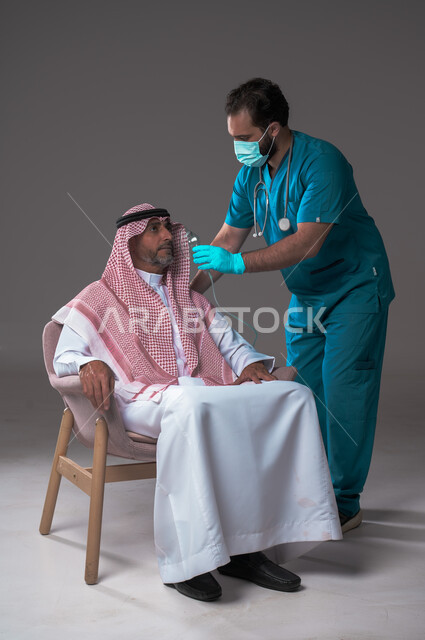اجراءات الفحص الطبي العامة لكبار السن، بورتريه لممرض عربي خليجي سعودي يلبس اليونيفورم والقفازات والكمامة الوقائية يقوم بفحص أذن مريض مسن يرتدي الثوب التقليدي والشماغ يجلس على كرسي، استخدام الأدوات الطبية الحديثة، خلفية رمادية