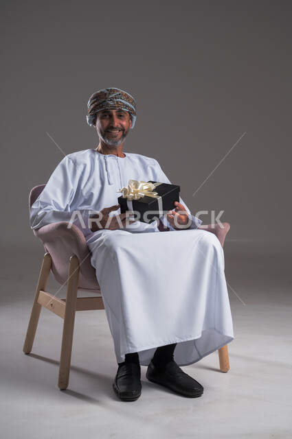  التهنئات والمباركات بالنجاح والأفراح, بورتريه لرجل عربي  خليجي عماني مبتسم يرتدي العمامة والدشداشة يجلس على كرسي مريح يحمل بيده علبة هدية، أعياد ومفاجآت ومناسبات سعيدة، النظر الى الكاميرا بايماءات السعادة والسرور، خلفية رمادية