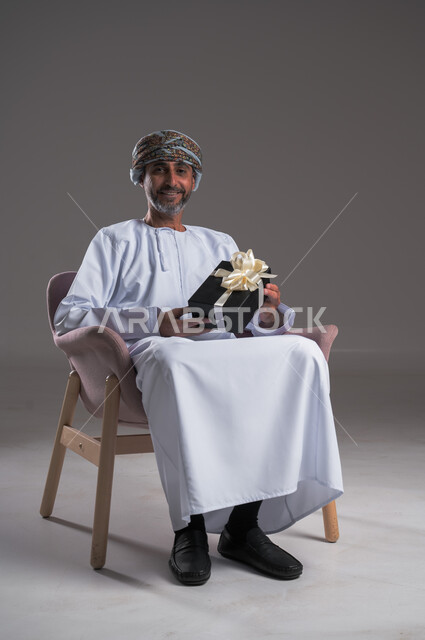  التهنئات والمباركات بالنجاح والأفراح, بورتريه لرجل عربي  خليجي عماني مبتسم يرتدي العمامة والدشداشة يجلس على كرسي مريح يحمل بيده علبة هدية، أعياد ومفاجآت ومناسبات سعيدة، النظر الى الكاميرا بايماءات السعادة والسرور، خلفية رمادية