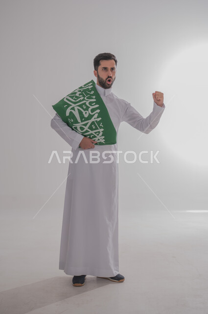 المشاركة في الاحتفال بالمناسبات والأعياد الوطنية، مفهوم الولاء وحب الوطن، بورتريه لشاب عربي خليجي سعودي يرتدي الثوب التقليدي واقف يضع علم المملكة على أكتافه يرفع يده بإيماءات الحماس والقوة، إحياء ذكرى اليوم الوطني السعودي، الجسم بالطول الكامل، خلفية رمادية