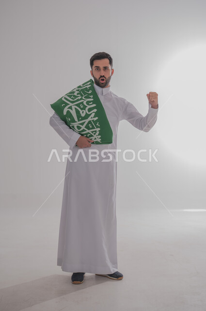 المشاركة في الاحتفال بالمناسبات والأعياد الوطنية، مفهوم الولاء وحب الوطن، بورتريه لشاب عربي خليجي سعودي يرتدي الثوب التقليدي واقف يضع علم المملكة على أكتافه يرفع يده بإيماءات الحماس والقوة، إحياء ذكرى اليوم الوطني السعودي، الجسم بالطول الكامل، خلفية رمادية