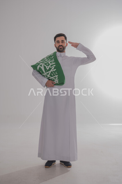 المشاركة في الاحتفال بالمناسبات والأعياد الوطنية، مفهوم الولاء وحب الوطن، بورتريه لشاب عربي خليجي سعودي يرتدي الثوب التقليدي واقف يضع علم المملكة على أكتافه يقف بشكل مستقيم ويقدم التحية، إحياء ذكرى اليوم الوطني السعودي، الجسم بالطول الكامل، خلفية رمادية