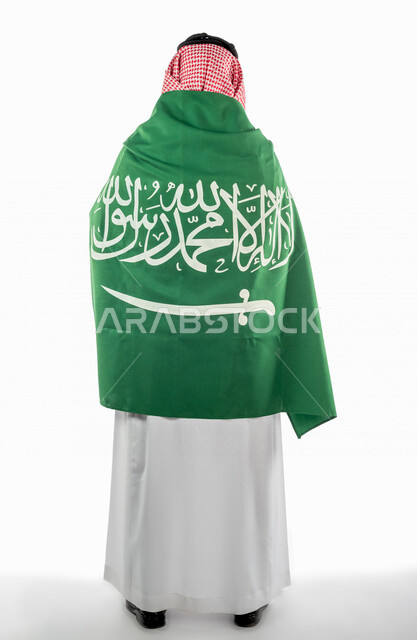 تصوير من الخلف لرجل عربي خليجي سعودي يضع على ظهره علم المملكة العربية السعودية ، ذكرى اليوم الوطني السعودي ،الاحتفال بالعيد الوطني السعودي