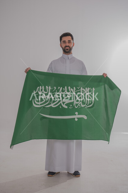 رفع علم المملكة العربية السعودية وإحياء ذكرى اليوم الوطني السعودي ٢٣ سبتمبر، تقديم الولاء والحب للوطن، الاحتفال بالمناسبات والأعياد الوطنية، بورتريه لشاب عربي خليجي سعودي يرتدي الثوب التقليدي يقف وينظر للكاميرا بسعادة وفخر، الجسم بالطول الكامل، خلفية رمادية