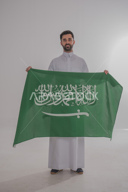 رفع علم المملكة العربية السعودية وإحياء ذكرى اليوم الوطني السعودي ٢٣ سبتمبر، تقديم الولاء والحب للوطن، الاحتفال بالمناسبات والأعياد الوطنية، بورتريه لشاب عربي خليجي سعودي يرتدي الثوب التقليدي يقف وينظر للكاميرا بسعادة وفخر، الجسم بالطول الكامل، خلفية رمادية