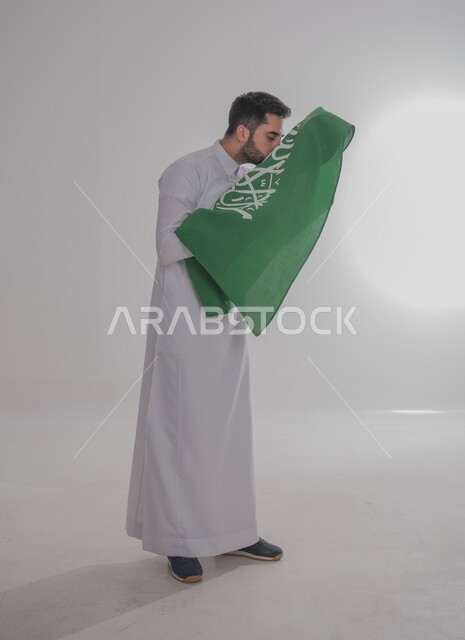 تقديم الولاء والحب للوطن، المشاركة في الاحتفال بالمناسبات والأعياد الوطنية، بورتريه لشاب عربي خليجي سعودي يرتدي الثوب التقليدي يقف ويحمل علم المملكة ويقوم بتقبليه، إحياء ذكرى اليوم الوطني السعودي ٢٣ سبتمبر، الجسم بالطول الكامل، خلفية رمادية
