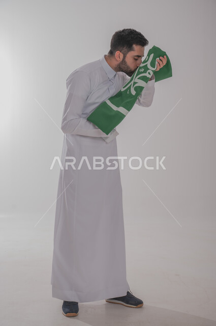 تقديم الولاء والحب للوطن، المشاركة في الاحتفال بالمناسبات والأعياد الوطنية، بورتريه لشاب عربي خليجي سعودي يرتدي الثوب التقليدي يقف ويحمل علم المملكة ويقوم بتقبليه، إحياء ذكرى اليوم الوطني السعودي ٢٣ سبتمبر، الجسم بالطول الكامل، خلفية رمادية