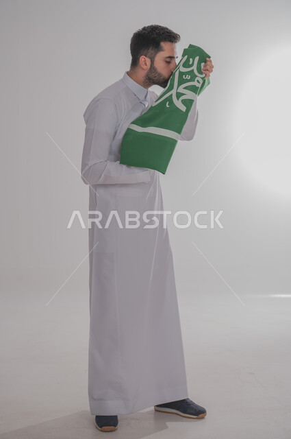 تقديم الولاء والحب للوطن، المشاركة في الاحتفال بالمناسبات والأعياد الوطنية، بورتريه لشاب عربي خليجي سعودي يرتدي الثوب التقليدي يقف ويحمل علم المملكة ويقوم بتقبليه، إحياء ذكرى اليوم الوطني السعودي ٢٣ سبتمبر، الجسم بالطول الكامل، خلفية رمادية