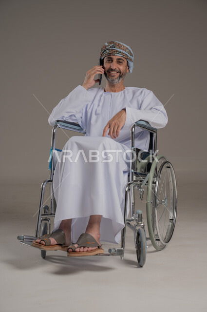  اجراء مكالمات هاتفية، ايماءات السرور والسعادة ،استخدام الاجهزة التقنية الحديثة، التواصل مع الاهل والاصدقاء، بورتريه لرجل عربي خليجي عماني مقعد يرتدي العمامة والدشداشة يجلس على كرسي متحرك يحمل بيده الهاتف المحمول، صورة لكامل الجسم، خلفية رمادية