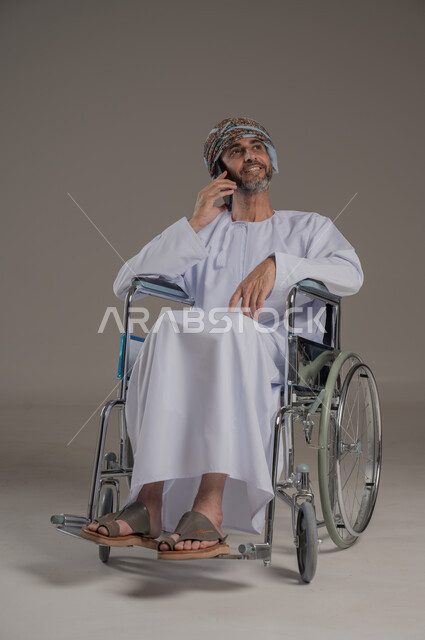 Making phone calls, gestures of pleasure and happiness, using modern technical devices, communicating with family and friends, portrait of a disabled Gulf Arab Omani man wearing a turban and dishdasha sitting in a wheelchair holding a mobile phone in his hand, full body image, gray background