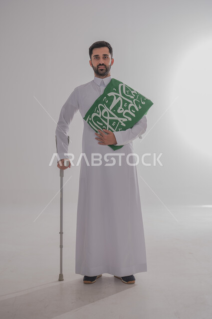 مشاركة ذوي الاحتياجات الخاصة بالاحتفال بالمناسبات والأعياد الوطنية، إحياء ذكرى اليوم الوطني السعودي، تقديم الولاء والحب للوطن، بورتريه لشاب عربي خليجي سعودي يرتدي الثوب التقليدي يستند إلى عكاز طبي ويحمل راية المملكة على كتفه، الجسم بالطول الكامل، خلفية رمادية