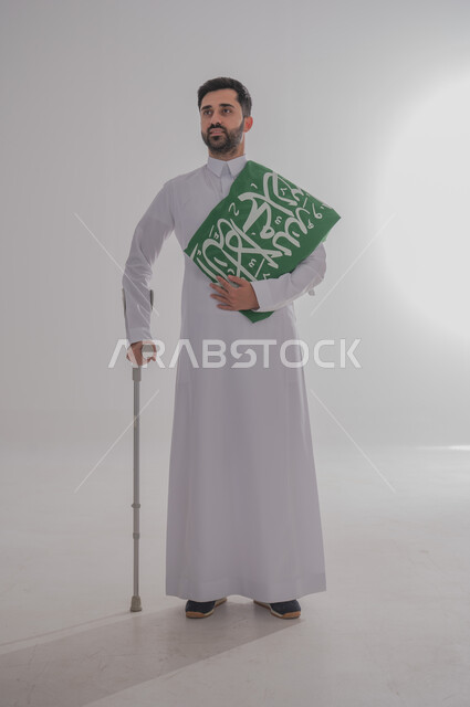 Participation of people with special needs in celebrating national occasions and holidays, commemorating Saudi National Day, presenting loyalty and love for the homeland, portrait of a young Saudi Gulf Arab man wearing traditional dress leaning on a medical crutch and carrying the Kingdom's flag on his shoulder, full body, gray background