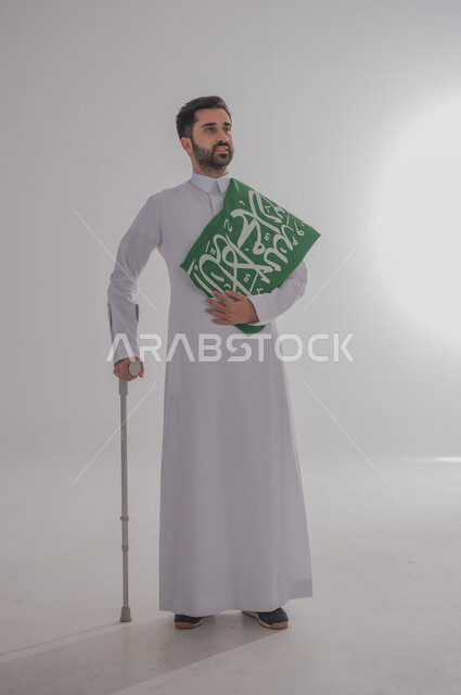 مشاركة ذوي الاحتياجات الخاصة بالاحتفال بالمناسبات والأعياد الوطنية، إحياء ذكرى اليوم الوطني السعودي، تقديم الولاء والحب للوطن، بورتريه لشاب عربي خليجي سعودي يرتدي الثوب التقليدي يستند إلى عكاز طبي ويحمل راية المملكة على كتفه، الجسم بالطول الكامل، خلفية رمادية