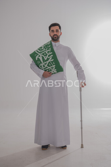 Participation of people with special needs in celebrating national occasions and holidays, commemorating Saudi National Day, presenting loyalty and love for the homeland, portrait of a young Saudi Gulf Arab man wearing traditional dress leaning on a medical crutch and carrying the Kingdom's flag on his shoulder, full body, gray background