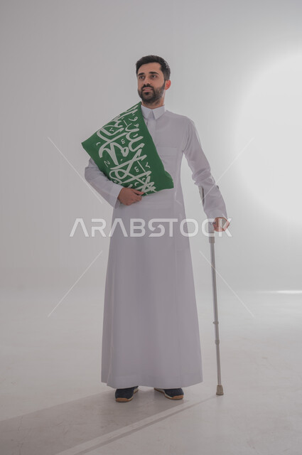 مشاركة ذوي الاحتياجات الخاصة بالاحتفال بالمناسبات والأعياد الوطنية، إحياء ذكرى اليوم الوطني السعودي، تقديم الولاء والحب للوطن، بورتريه لشاب عربي خليجي سعودي يرتدي الثوب التقليدي يستند إلى عكاز طبي ويحمل راية المملكة على كتفه، الجسم بالطول الكامل، خلفية رمادية