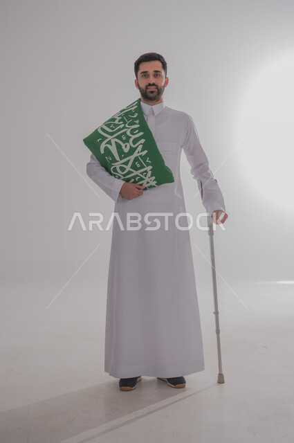 رفع علم المملكة العربية السعودية وإحياء ذكرى اليوم الوطني السعودي، تقديم الولاء والحب للوطن، الاحتفال بالمناسبات والأعياد الوطنية، بورتريه لشاب من ذوي الاحتياجات الخاصة عربي خليجي سعودي يرتدي الثوب التقليدي يستند إلى عكازه، الجسم بالطول الكامل، خلفية رمادية