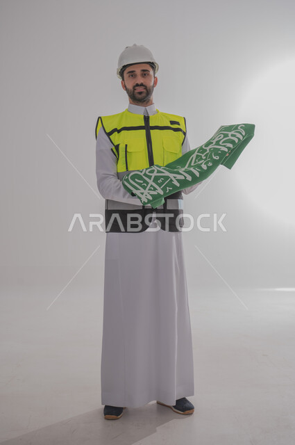 المشاركة بالاحتفال بالمناسبات والأعياد الوطنية، إحياء ذكرى اليوم الوطني السعودي، بورتريه لمهندس عربي خليجي سعودي يرتدي الثوب التقليدي والسترة وقبعة الحماية ويحمل راية المملكة العربية السعودية بين يديه بإيماءات الفخر والاعتزاز، الجسم بالطول الكامل، خلفية رمادية