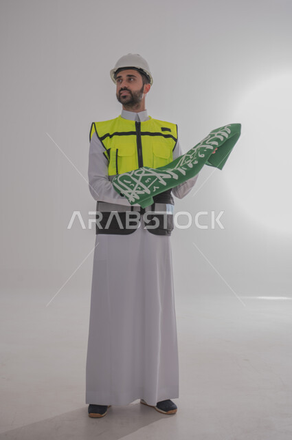 المشاركة بالاحتفال بالمناسبات والأعياد الوطنية، إحياء ذكرى اليوم الوطني السعودي، بورتريه لمهندس عربي خليجي سعودي يرتدي الثوب التقليدي والسترة وقبعة الحماية ويحمل راية المملكة العربية السعودية بين يديه بإيماءات الفخر والاعتزاز، الجسم بالطول الكامل، خلفية رمادية